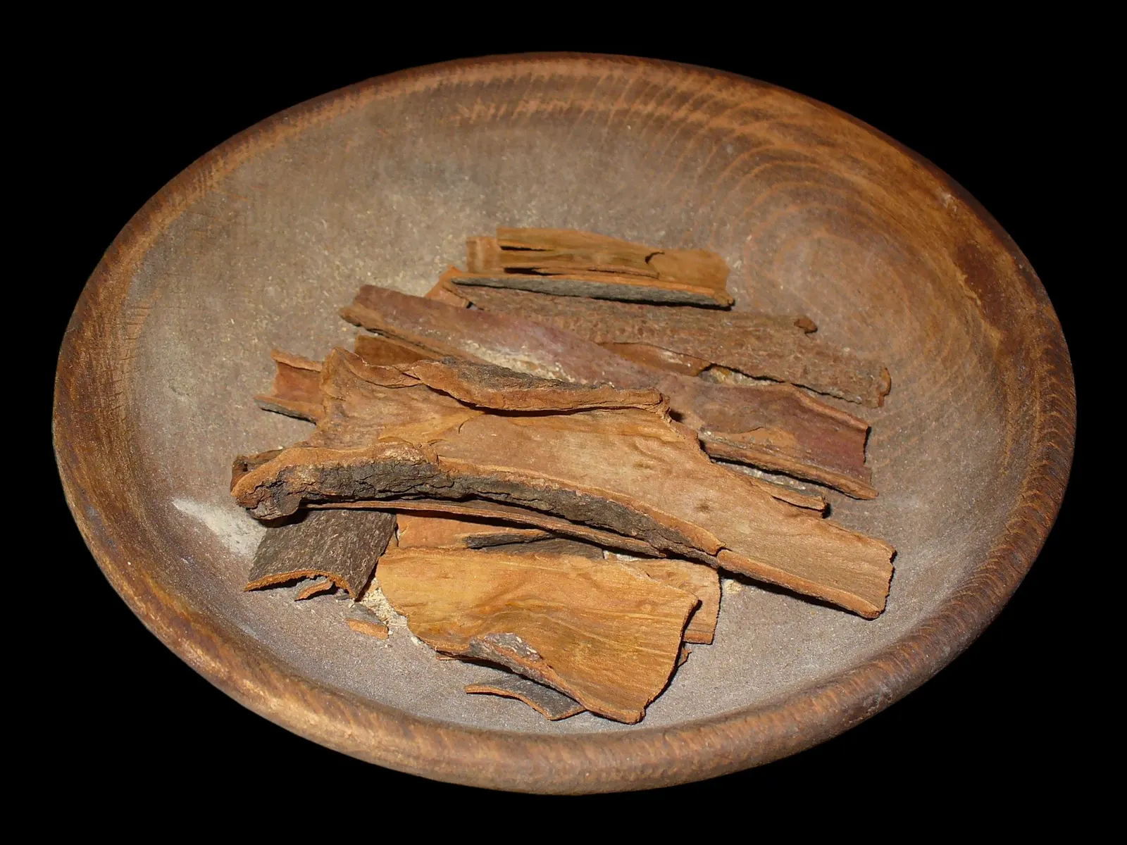Flat and curved pieces of reddish-brown cassia bark heaped in a wooden bowl on a black background, some edged with rough grey outer bark