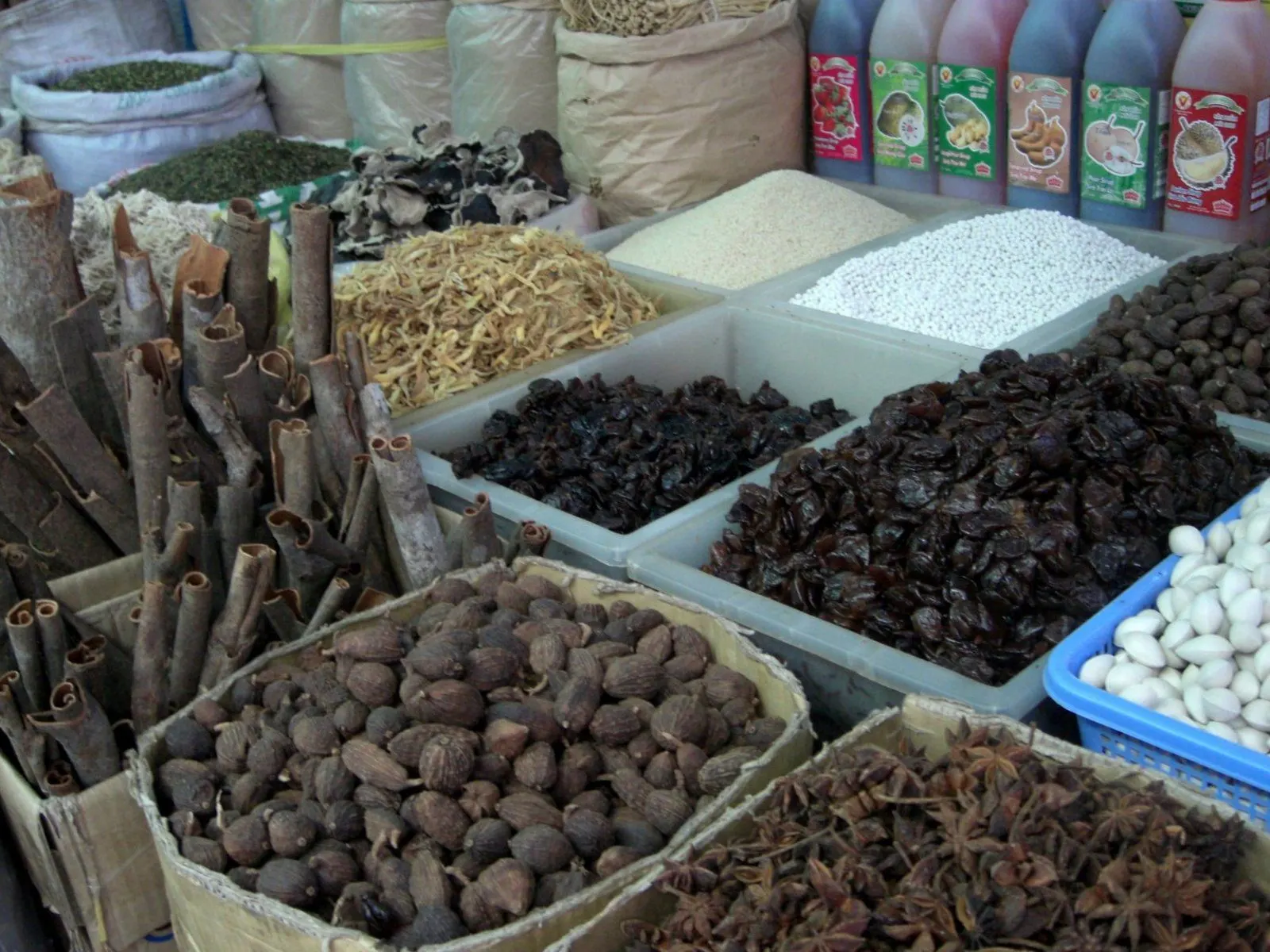 A market stall with upright cinnamon quills on the left, a box of whole nutmegs, a box of star anise, and trays of other dried goods behind