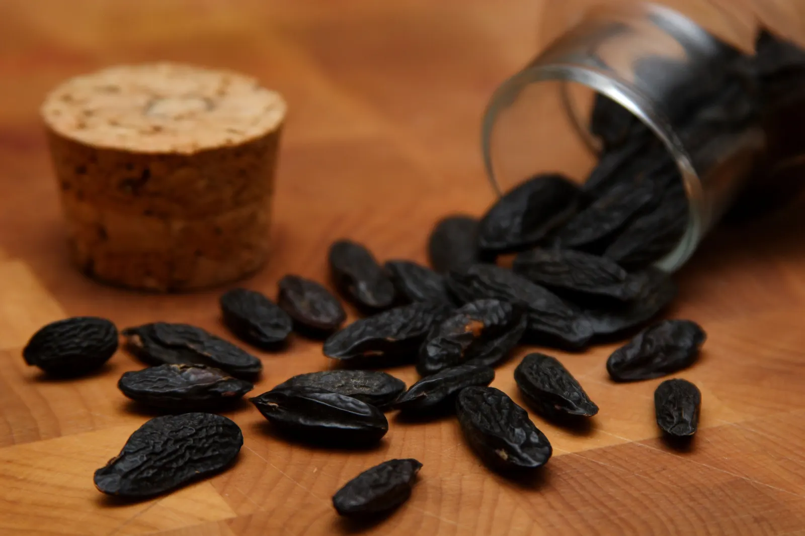 Black, wrinkled, almond-shaped tonka beans spilling from a tipped glass jar onto a wooden board, with a cork beside them